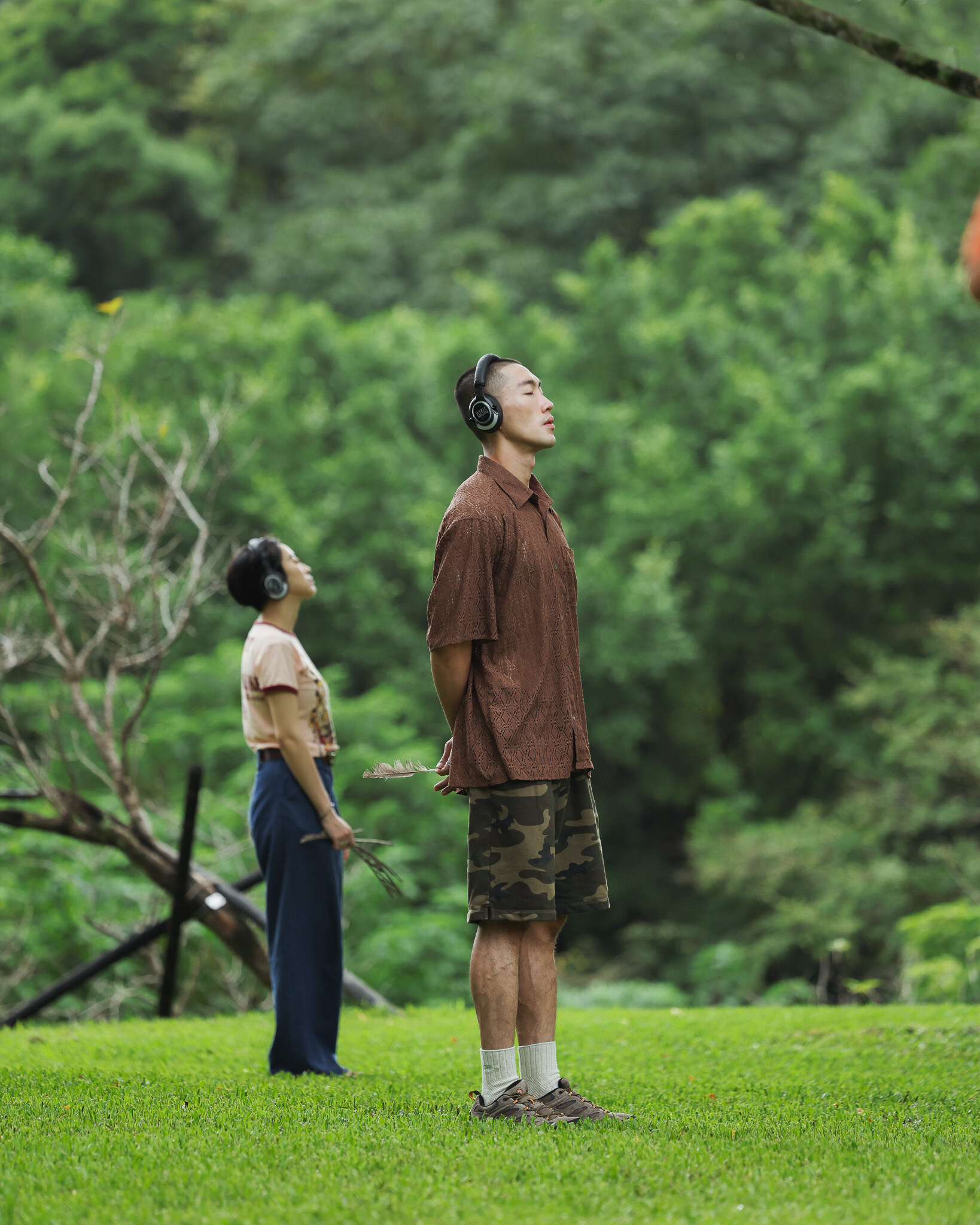 Two participants in headphones standing on a lawn in front of green forested hills during the Merrell sensory brand experience in Nangang Park, one with eyes closed and head tilted back